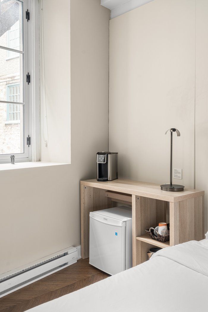A hotel room corner with a window, mini-fridge, coffee maker, lamp, and wooden shelf.