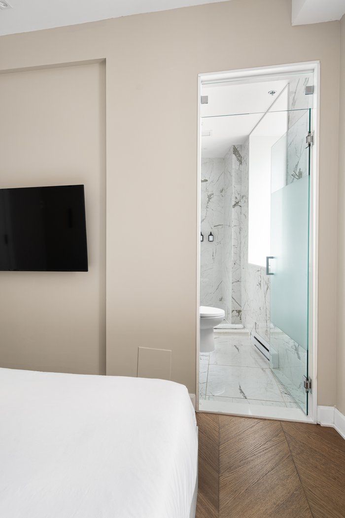 Bedroom with open glass door to bathroom. White bed, TV on wall, marble-tiled bathroom.