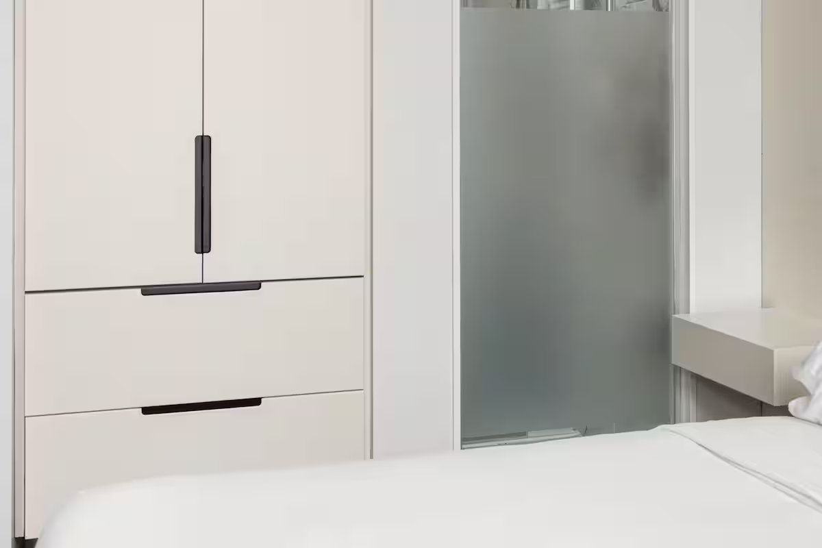 White bedroom closet and drawers with black handles, next to a bed and frosted glass shower door.