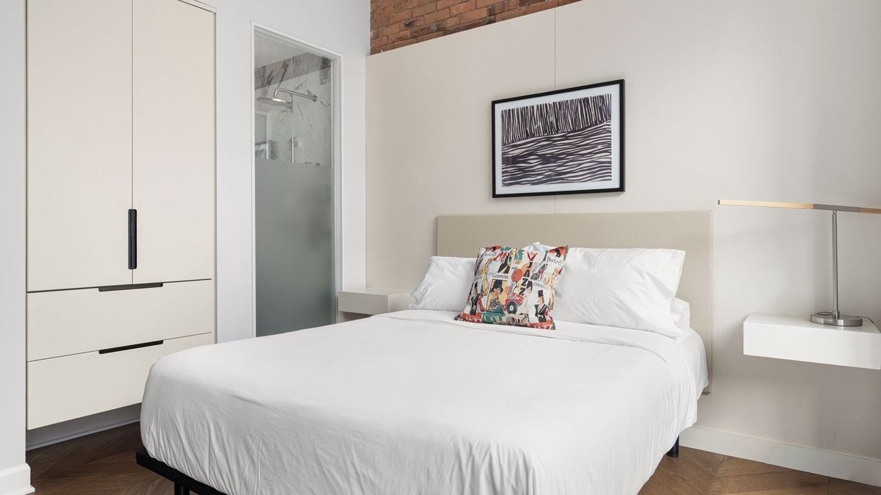 Bedroom with white bedding, headboard, and built-in closet. Artwork hangs above bed, with a frosted glass shower visible.