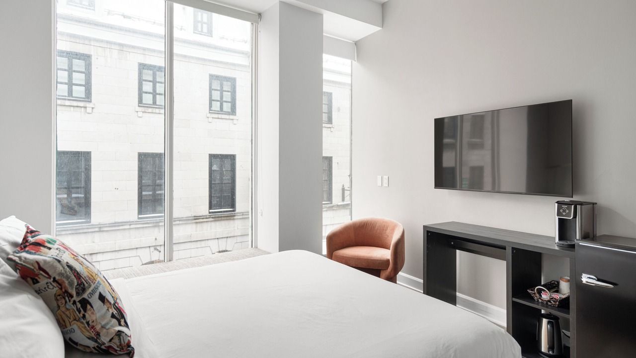 Bright bedroom with large window overlooking an old building, bed, TV, chair, and mini-fridge.