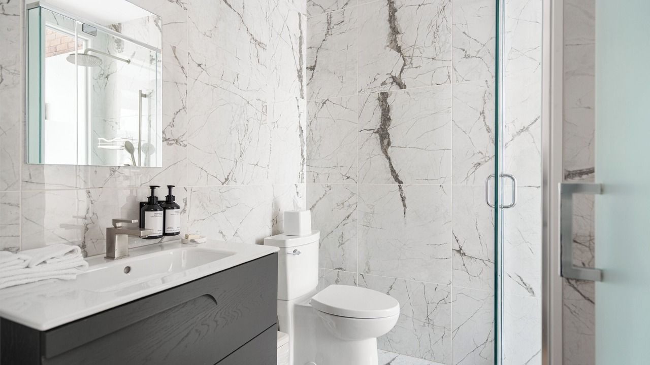 Bathroom with white marble tile walls, a dark gray vanity, toilet, and glass shower.