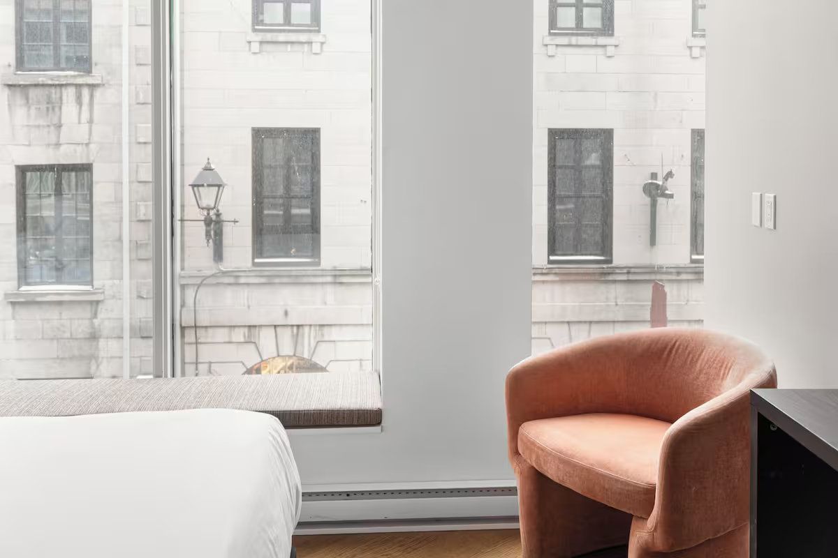 Room with peach armchair, window overlooking a building facade, and part of a bed.