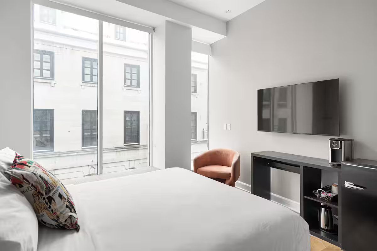 Bedroom with large window overlooking a street, white bed, chair, TV, and mini-fridge.