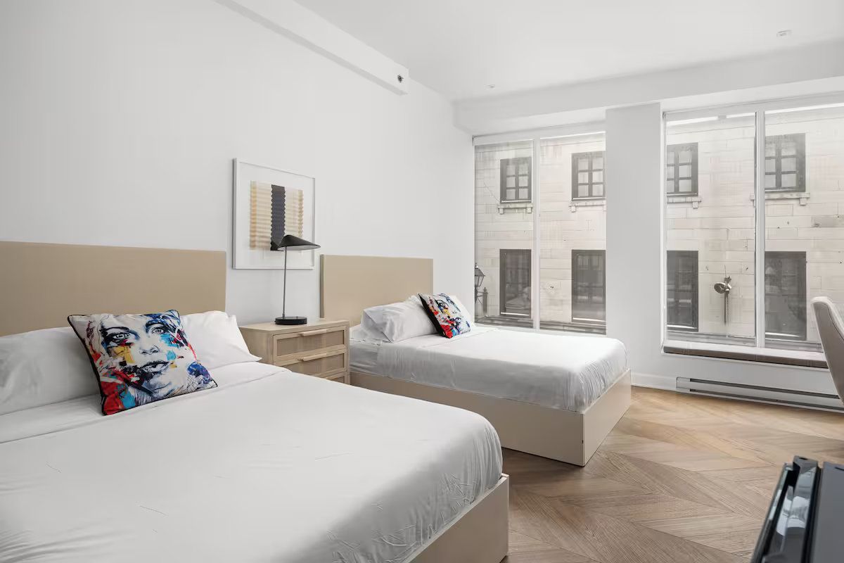 Two beds in a bright room with a large window overlooking a building.