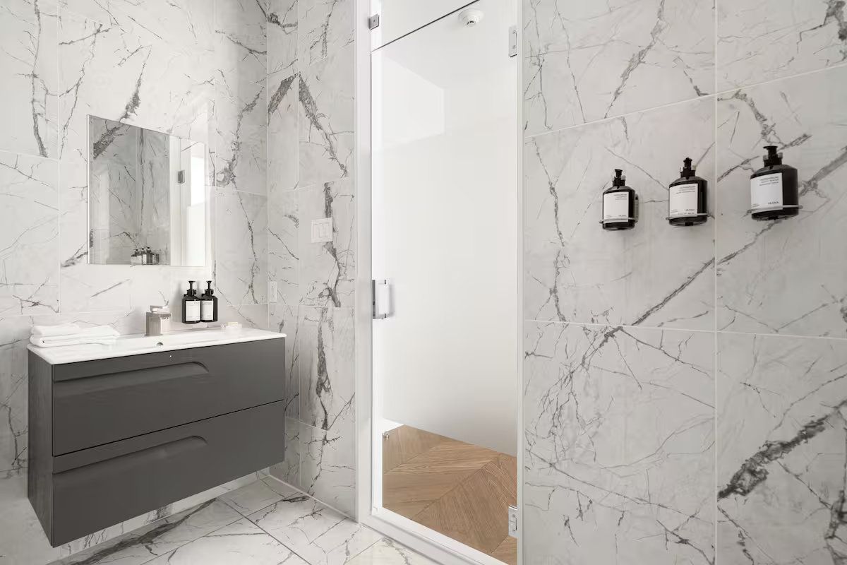 Modern bathroom with marble walls, gray vanity, and glass shower. Three black soap dispensers.