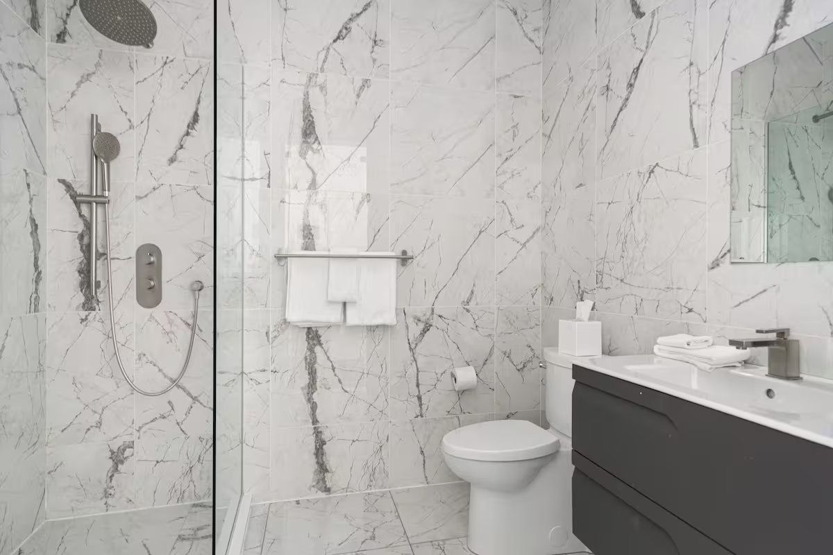 Modern bathroom with marble-look tiles, glass shower, toilet, dark gray vanity, and mirror.