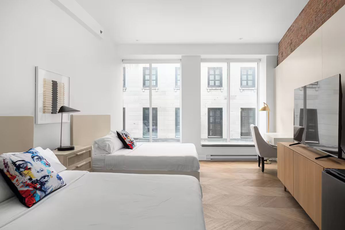 Hotel room with two beds, a desk, and large windows looking out onto a city street.