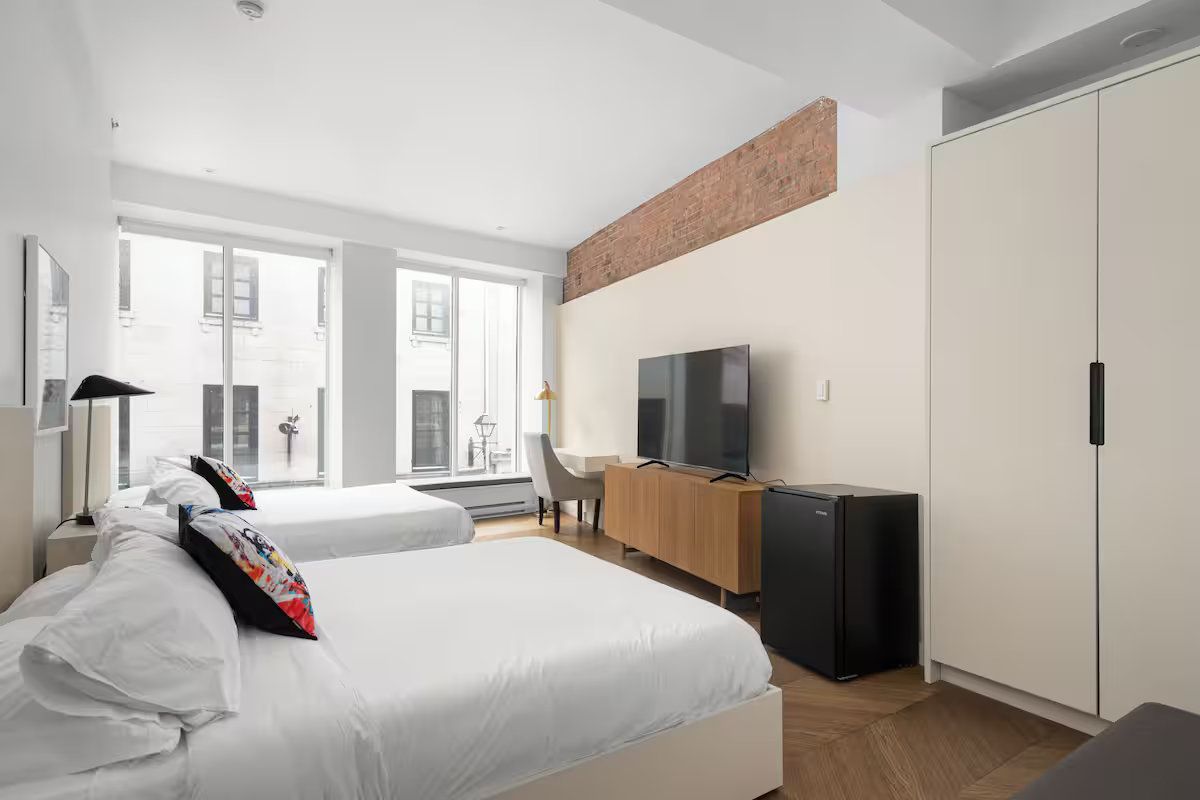 Modern hotel room with two beds, large windows, and a TV on a wooden console.