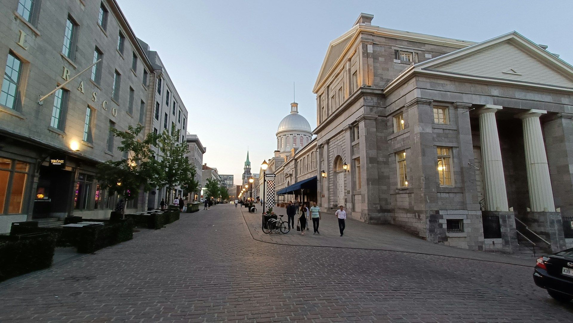 Cobblestone street lined with stone buildings, people, and a domed structure in the distance.