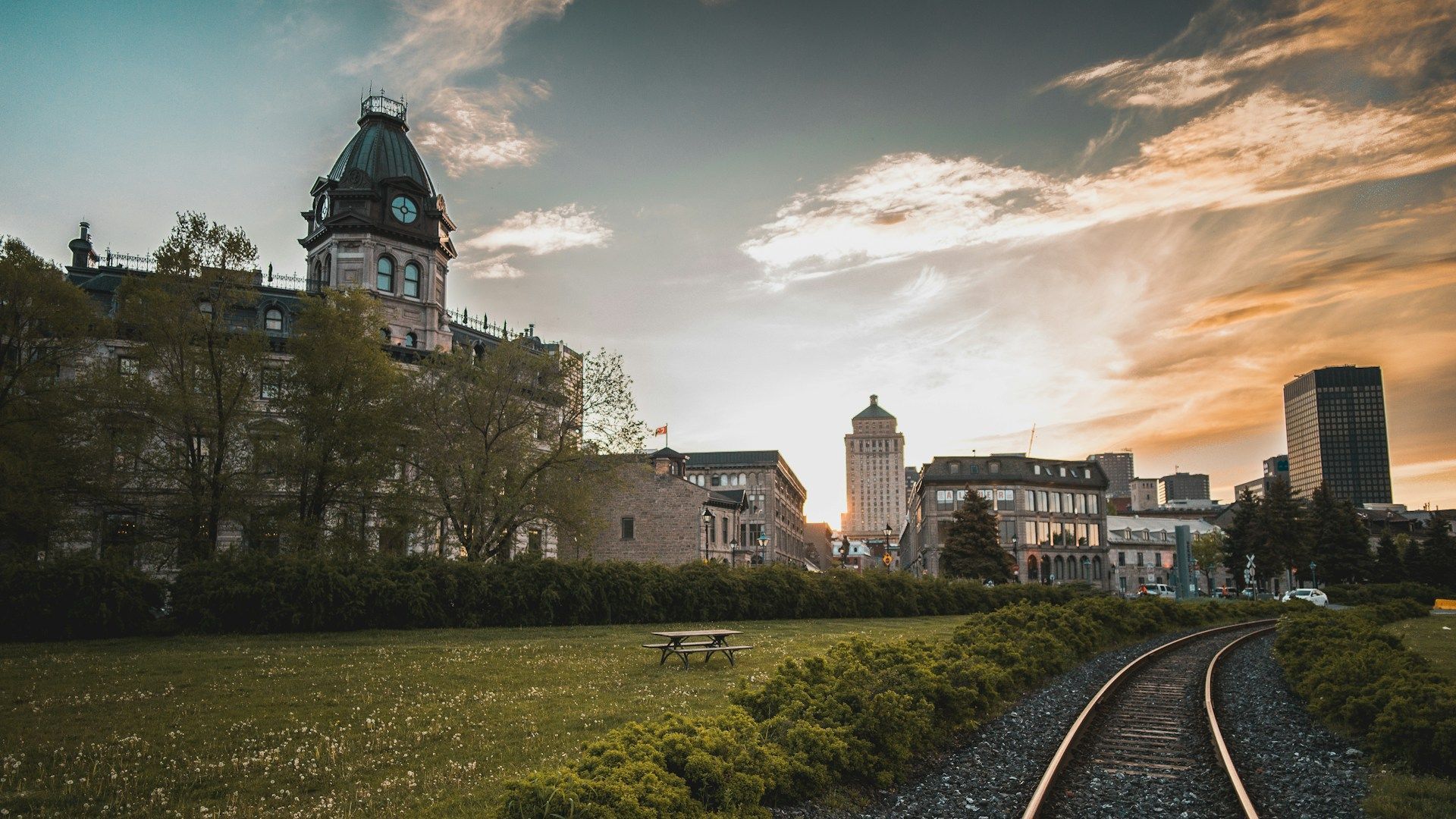 City skyline with clock tower and train tracks against a sunset sky.