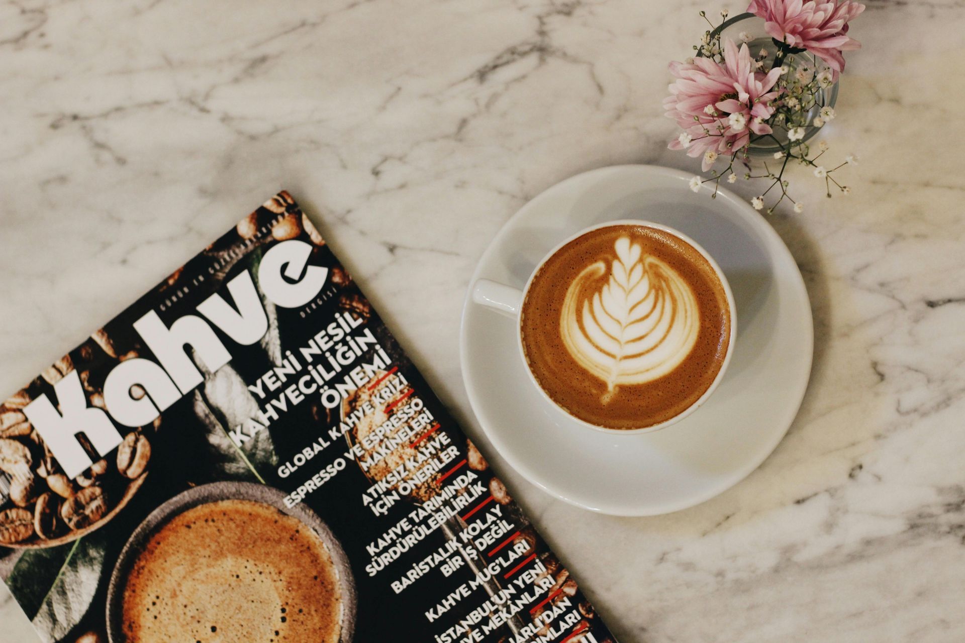 Latte with latte art on a saucer, next to a coffee magazine and a small floral arrangement, on a marble table.