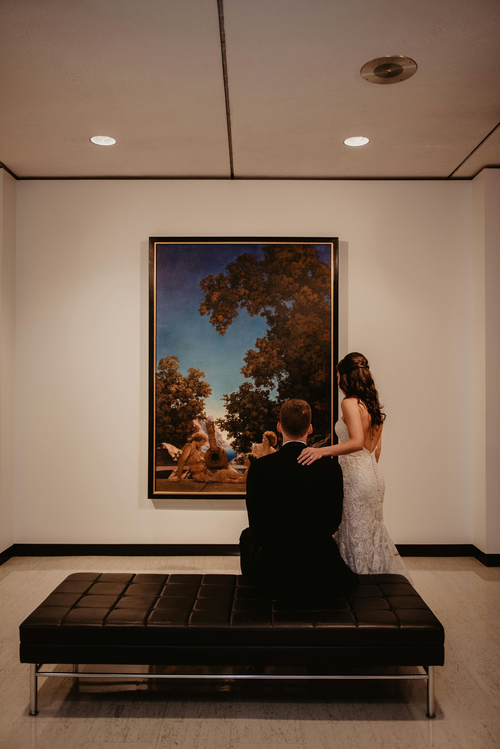 Couple on a bench, looking at a painting in an art gallery. The woman's arm is on the man's shoulder.