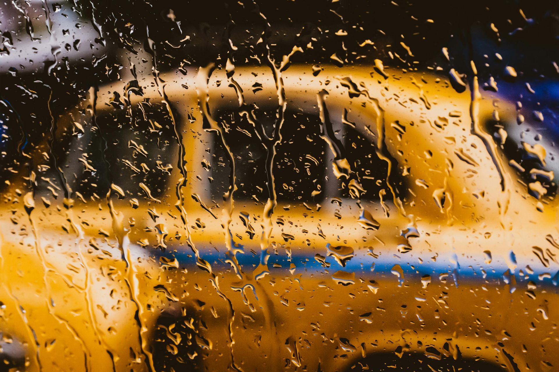 Yellow car blurred behind rain-streaked window; wet, close-up shot, golden hues.