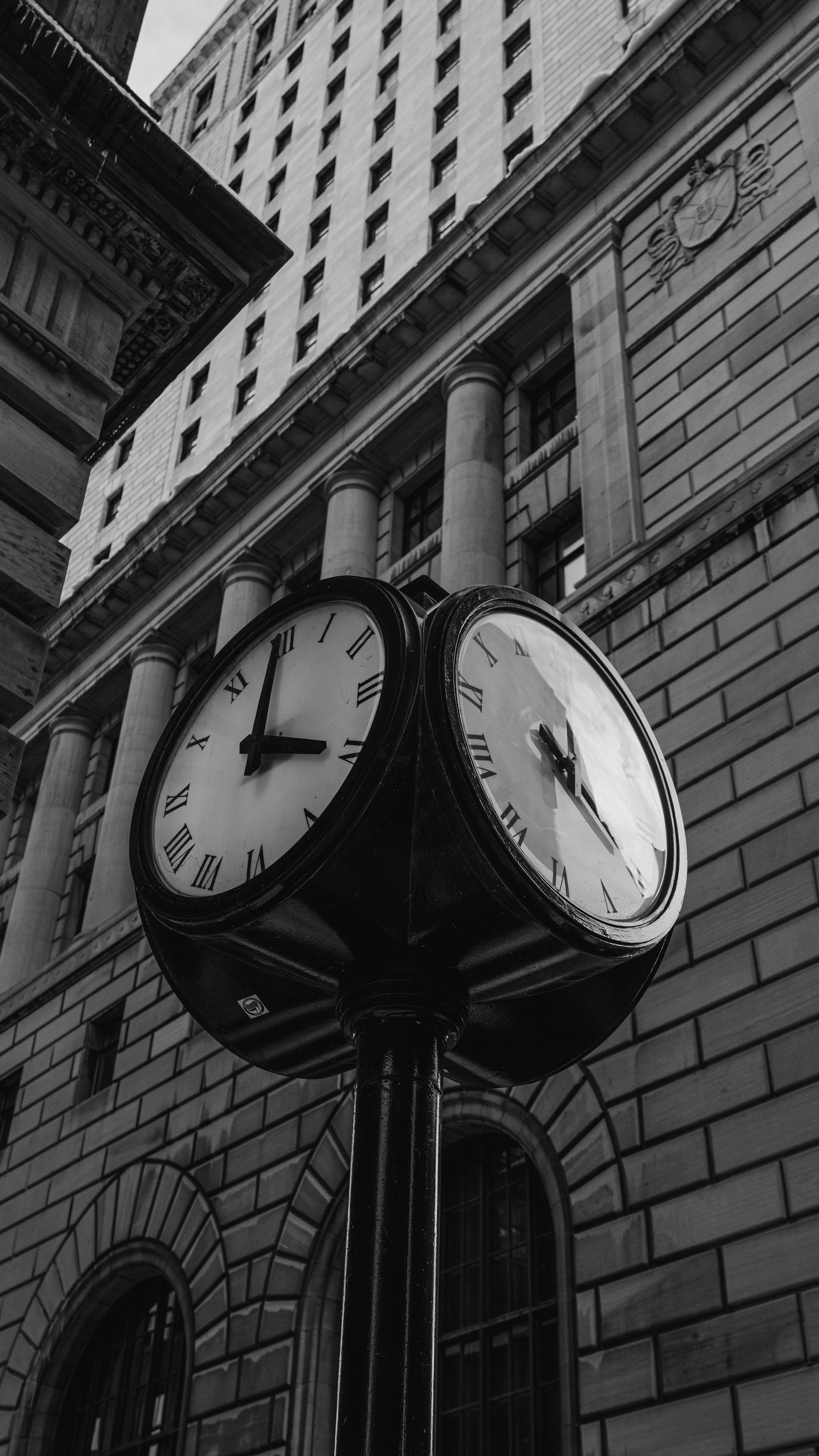Black and white street clock with two faces, in front of a tall brick building. One clock face reads nearly 1 o'clock.