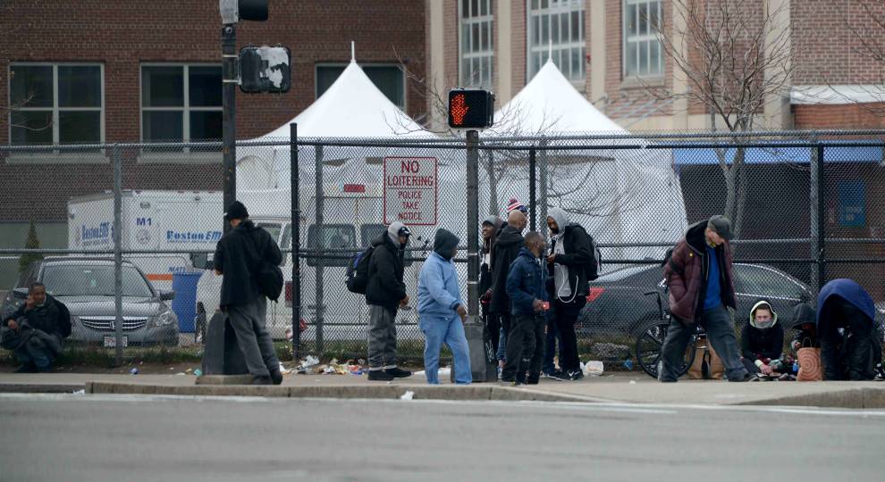 People gather near tents behind a fence on a city street. Some stand, some sit, near a crosswalk.