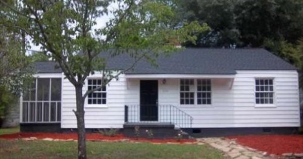 White one-story house with black roof, screened porch, and front door. Small tree in front.