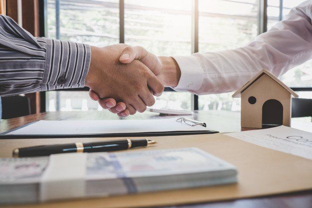 Two people shaking hands over a table with documents, money, and a small house model.
