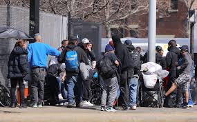 A group of people standing on a sidewalk near a building, some with bags and belongings.