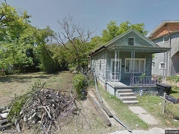 Weathered, light-green house with porch and steps, overgrown vegetation in front yard, and wood debris.