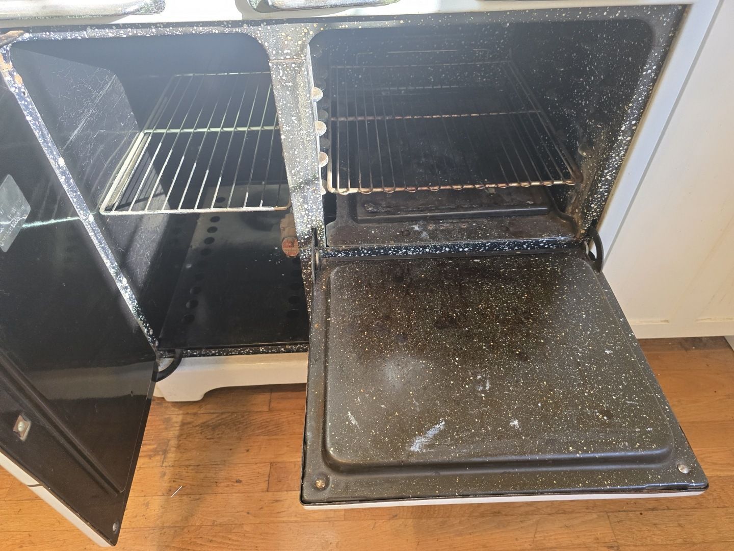 A dirty oven with the door open is sitting on a wooden floor.