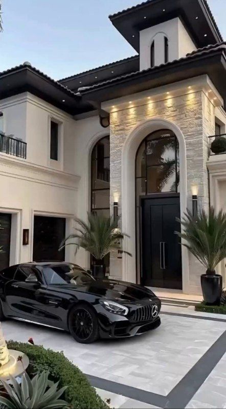 Black luxury car parked in front of a modern white mansion with a black door and arched entryway.