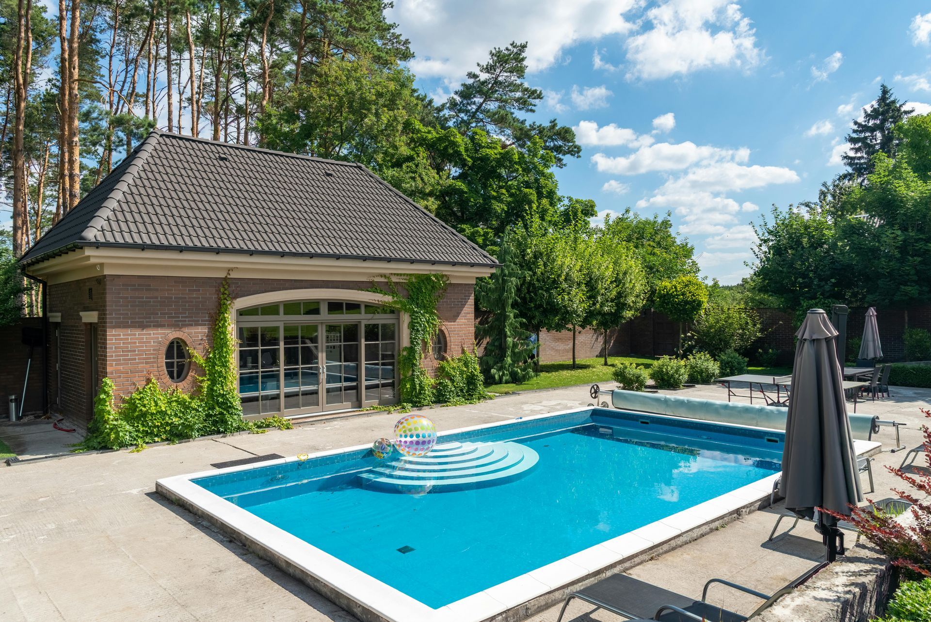 Pool with turquoise water, surrounded by concrete patio and brick building with glass windows. Trees in background.