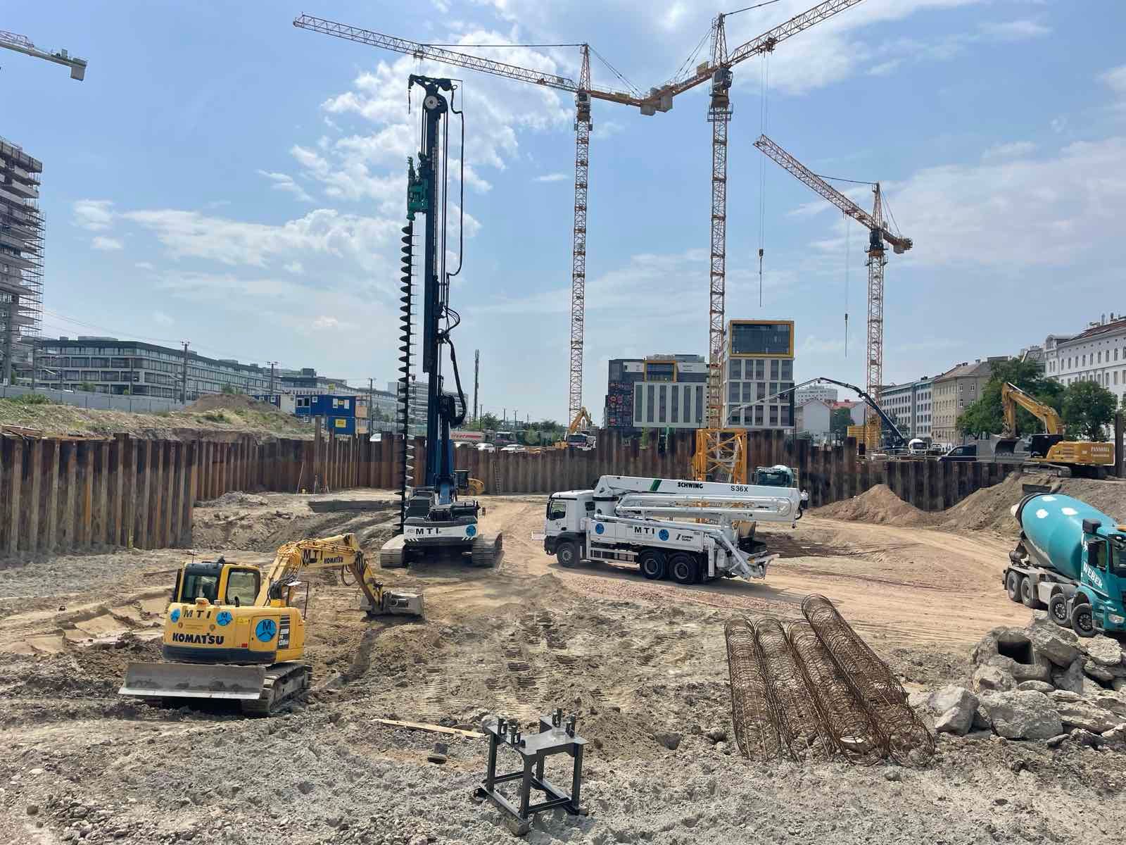 Baustelle mit schweren Maschinen, darunter Kräne, Bagger und Lastwagen, unter einem strahlenden Himmel.