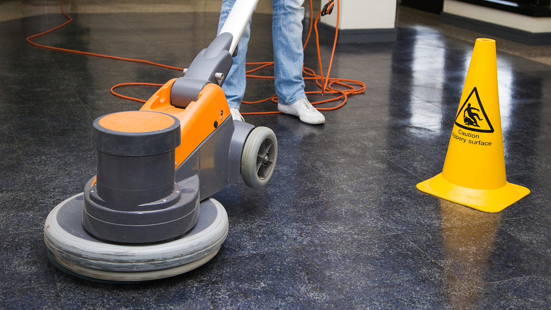 Person using an orange floor polisher in a room, next to a yellow caution cone.