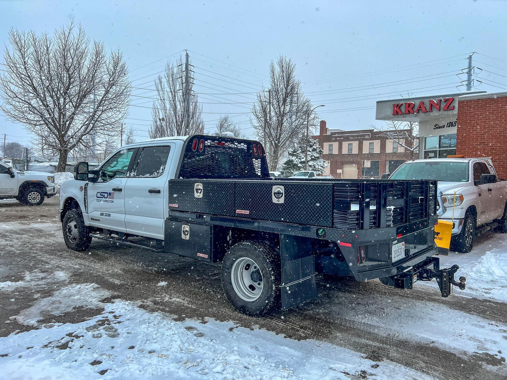 White work truck with a flatbed and toolboxes in a snowy parking lot.