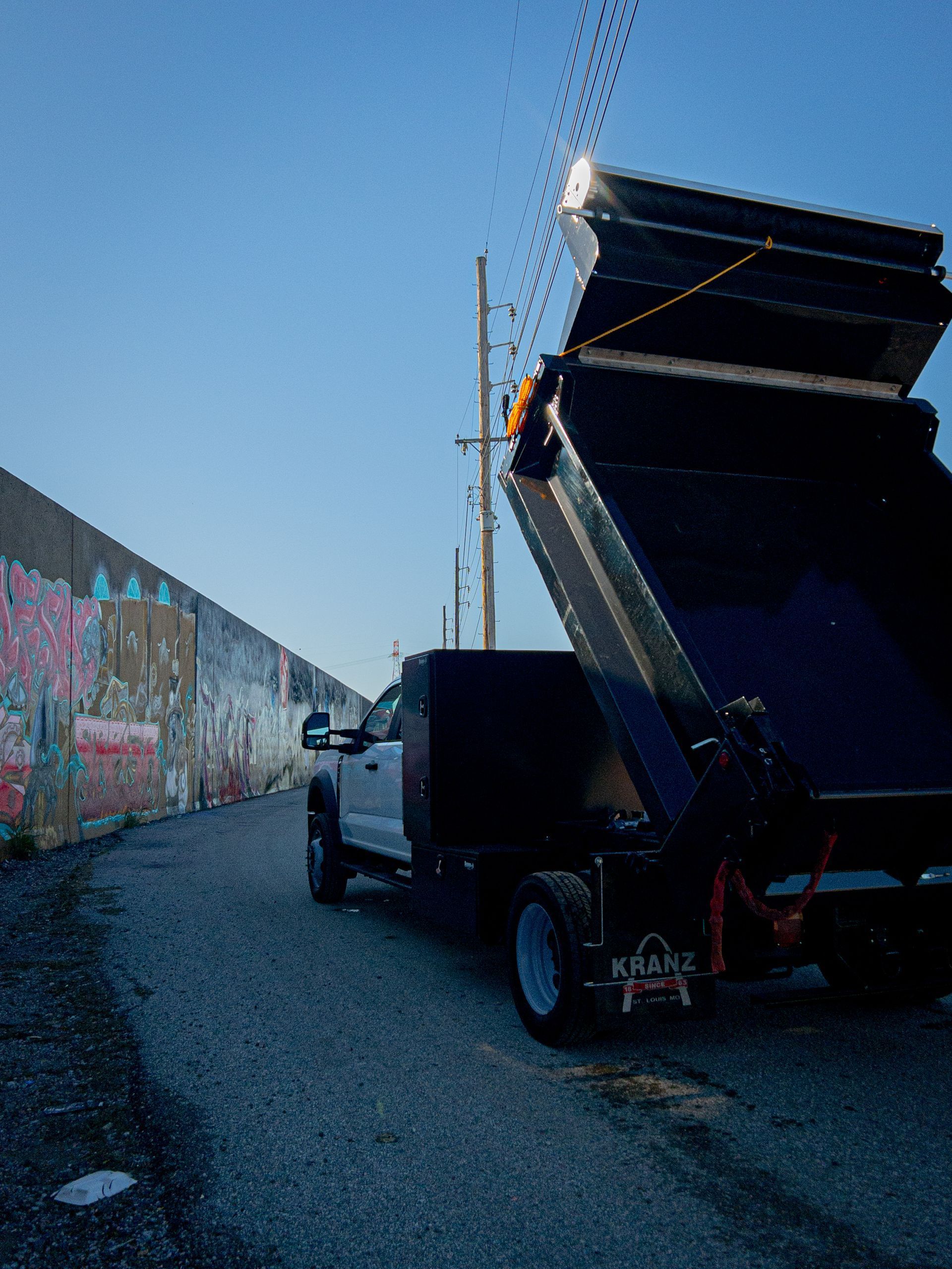 Dump truck with bed raised, parked on a paved alley next to a graffiti-covered wall.