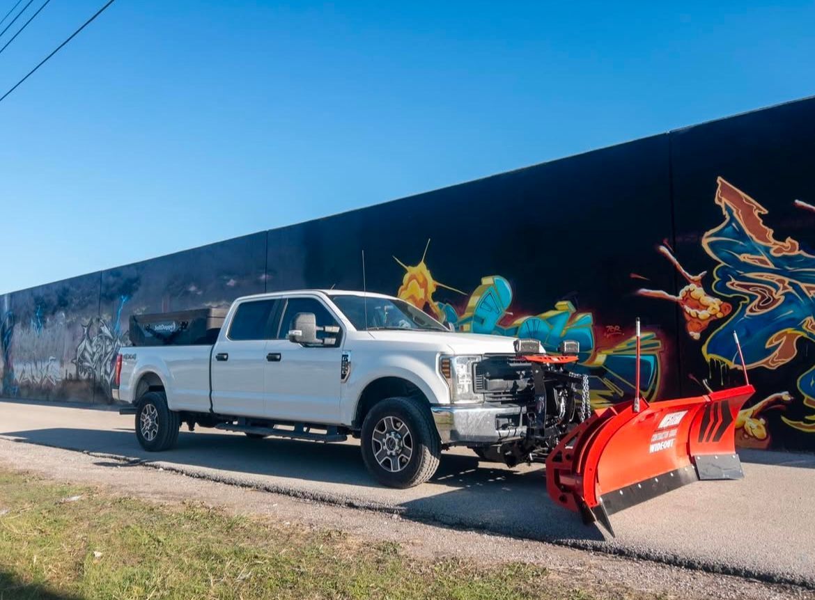 White pickup truck with snowplow parked on a road next to a mural.