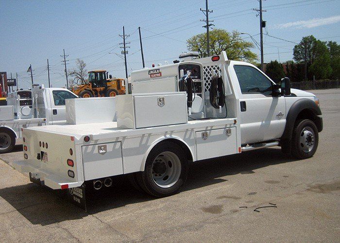 White service truck parked outdoors on a sunny day.  A flatbed with storage compartments and equipment is mounted on the truck.