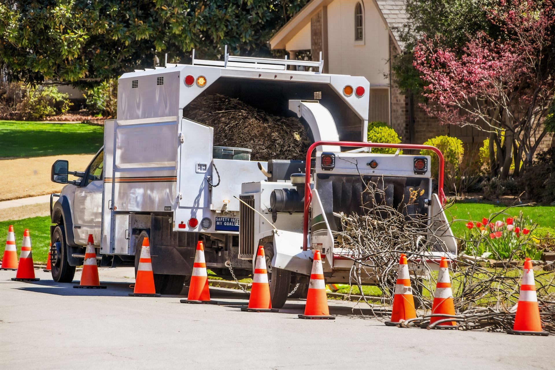 A wood chipper truck parked on a street, surrounded by traffic cones, processing tree branches.