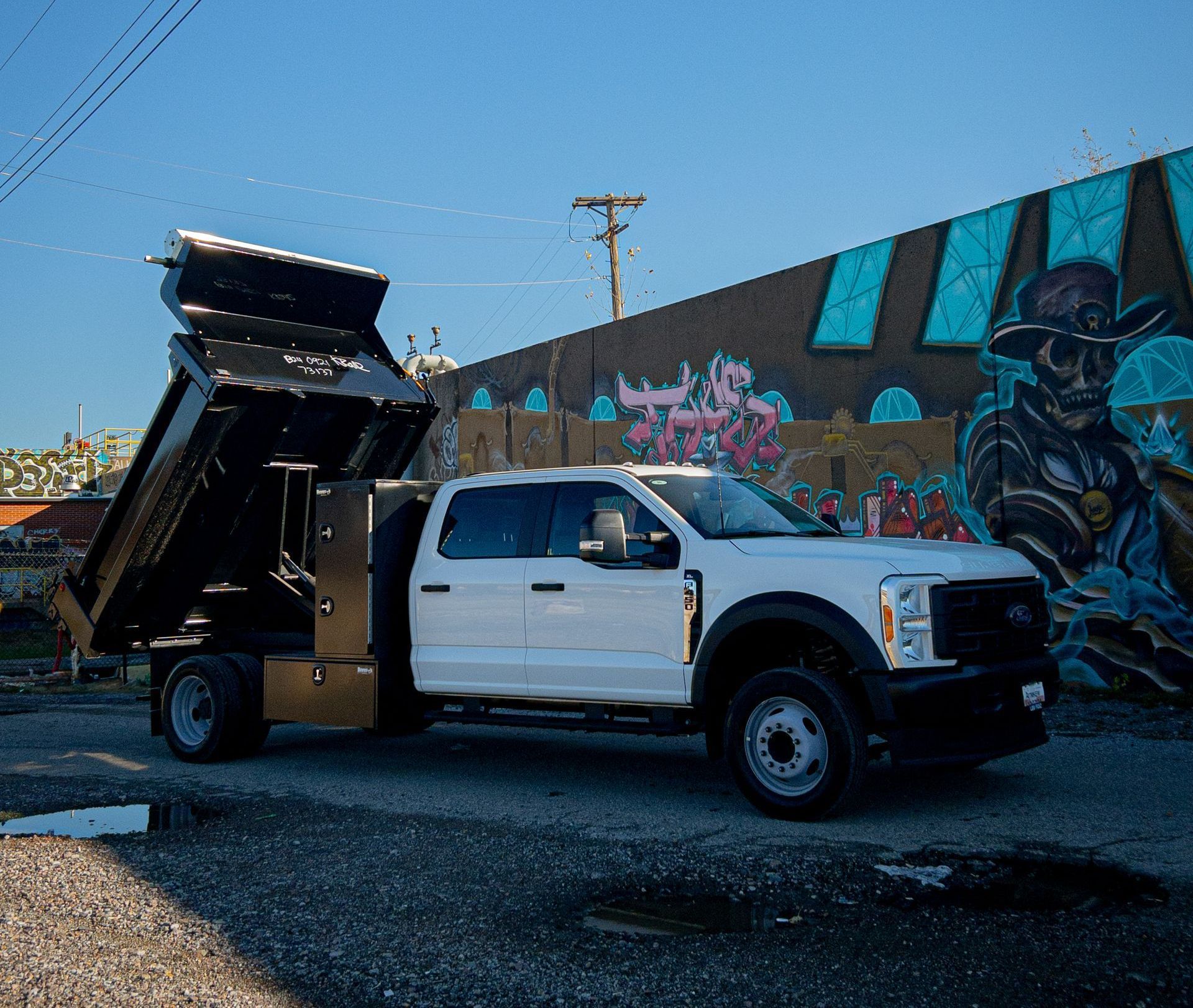 White dump truck with bed raised, parked in front of a graffiti wall.