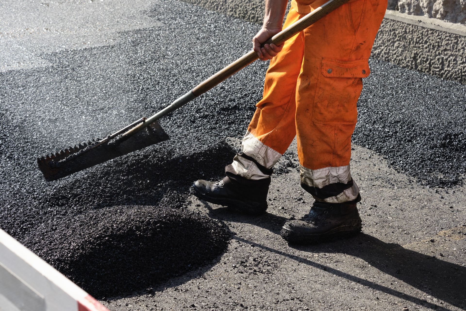 Construction worker spreads hot asphalt with a rake, leveling fresh pavement.