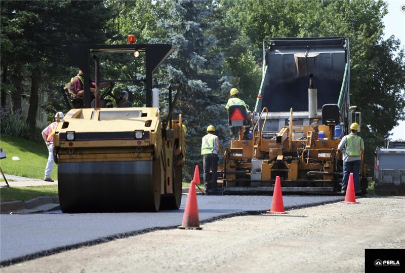 Road paving: Asphalt laid by a machine, workers in vests, roller compacting. Orange cones in street.