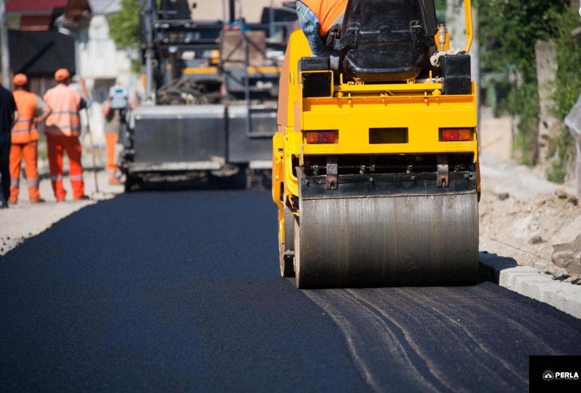Road paving in progress, yellow roller compacting fresh asphalt, workers in orange vests nearby.