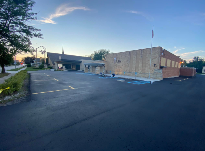 A church building with a parking lot and a flagpole under a blue and orange sky.