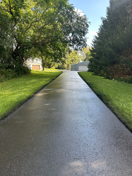 Newly paved driveway with wet, shiny surface. Green grass on either side, trees in background.