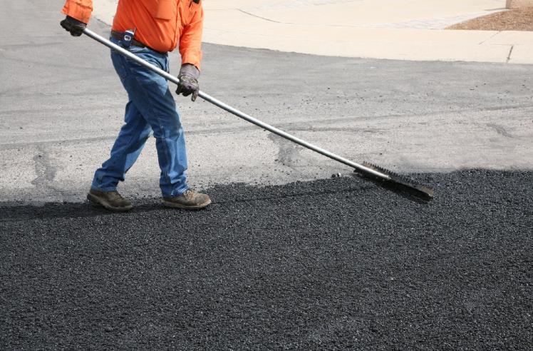 Person raking fresh asphalt on a road, wearing work clothes.