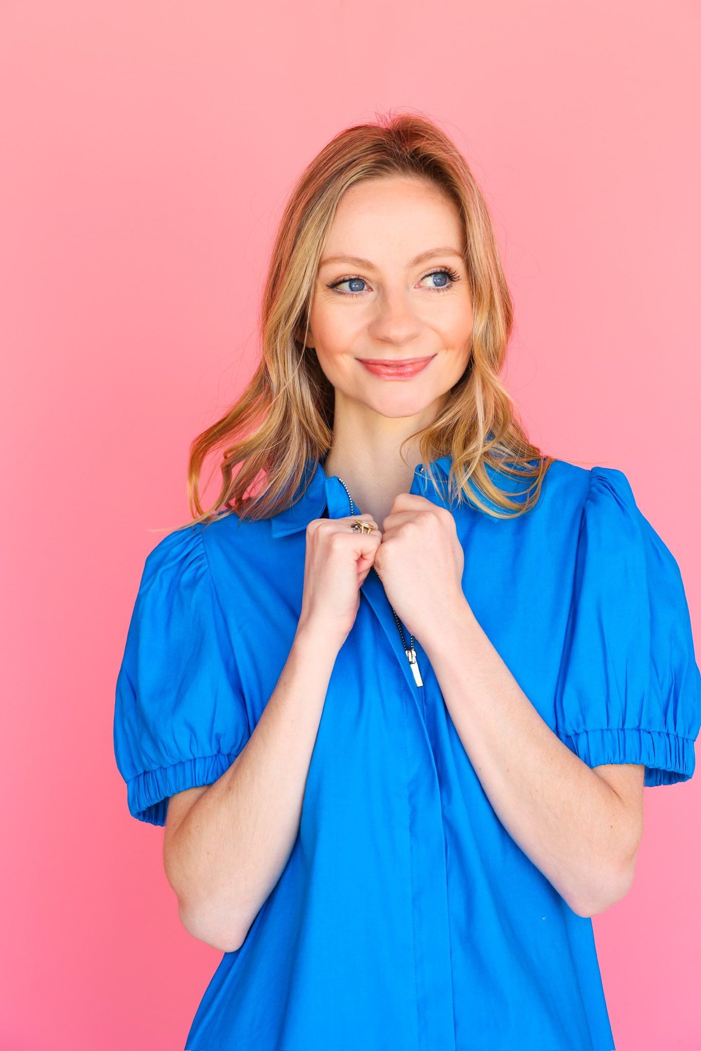 Woman in blue dress against a pink background, smiling, holding dress closed.
