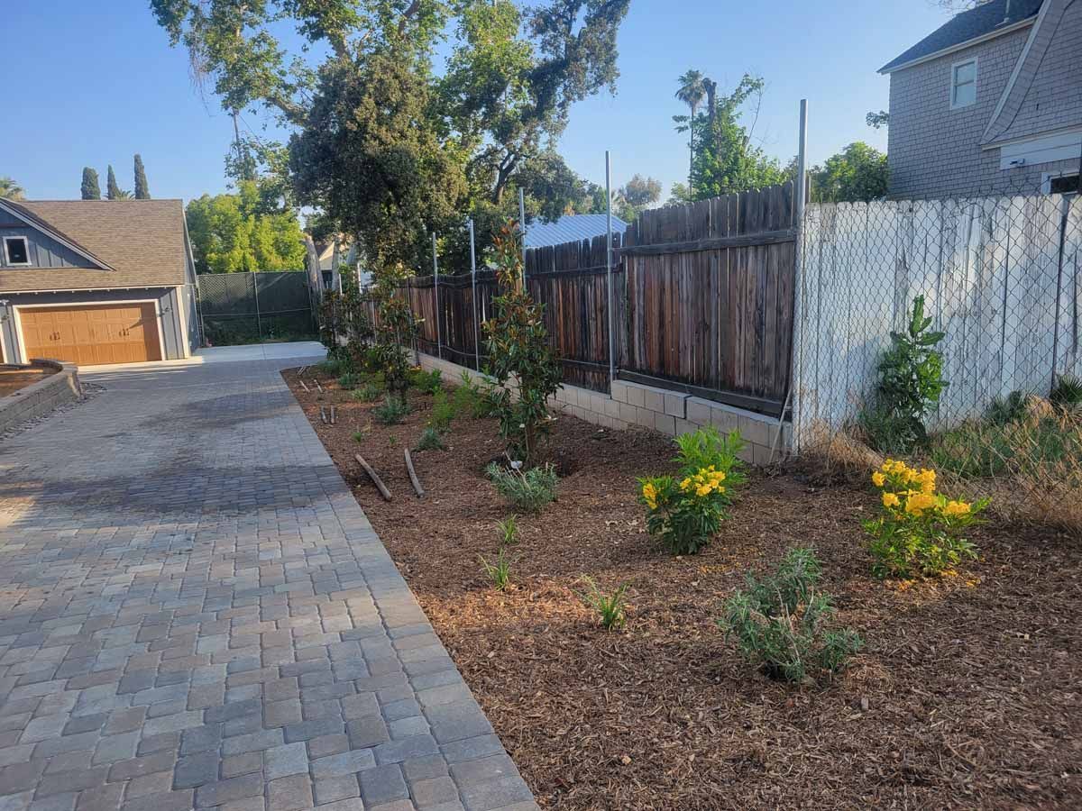 Driveway with brick pavers, wood fence, and landscaped beds with mulch and plants.