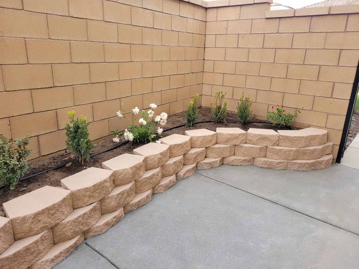A brown stone retaining wall with plants in a small garden, set against a light-brown brick wall.