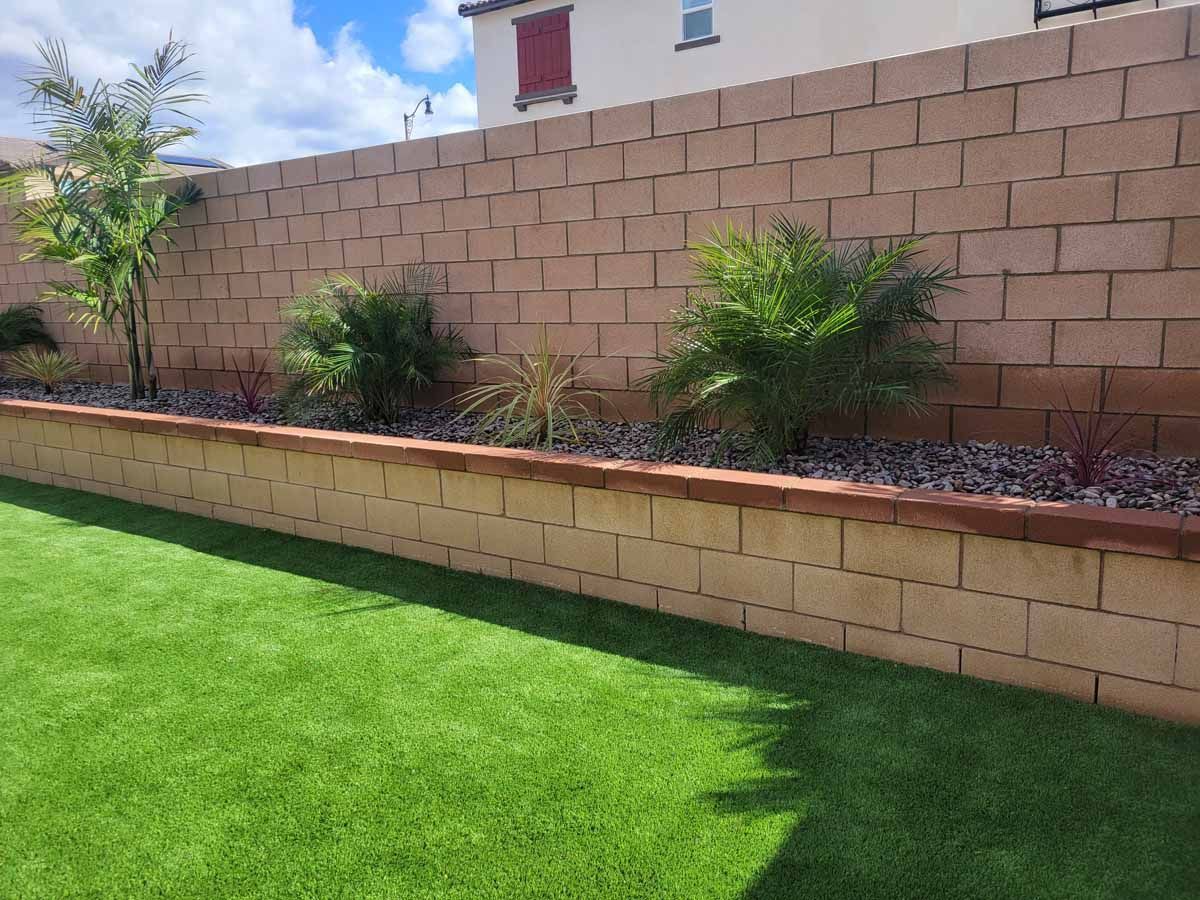 Green artificial lawn with tan block wall and raised garden bed, under blue sky.