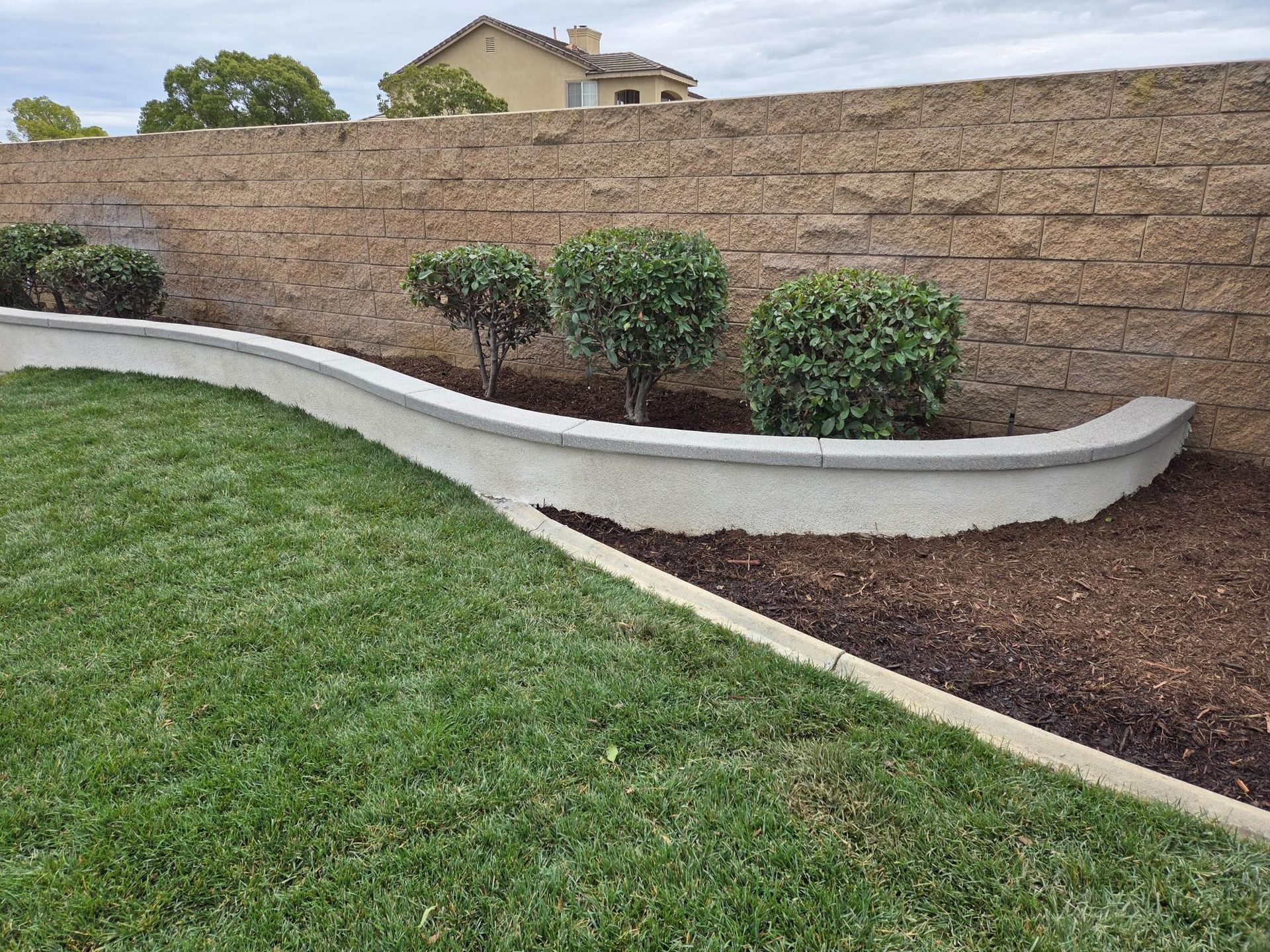 Curved retaining wall with trimmed bushes, green grass, and a brick wall backdrop.