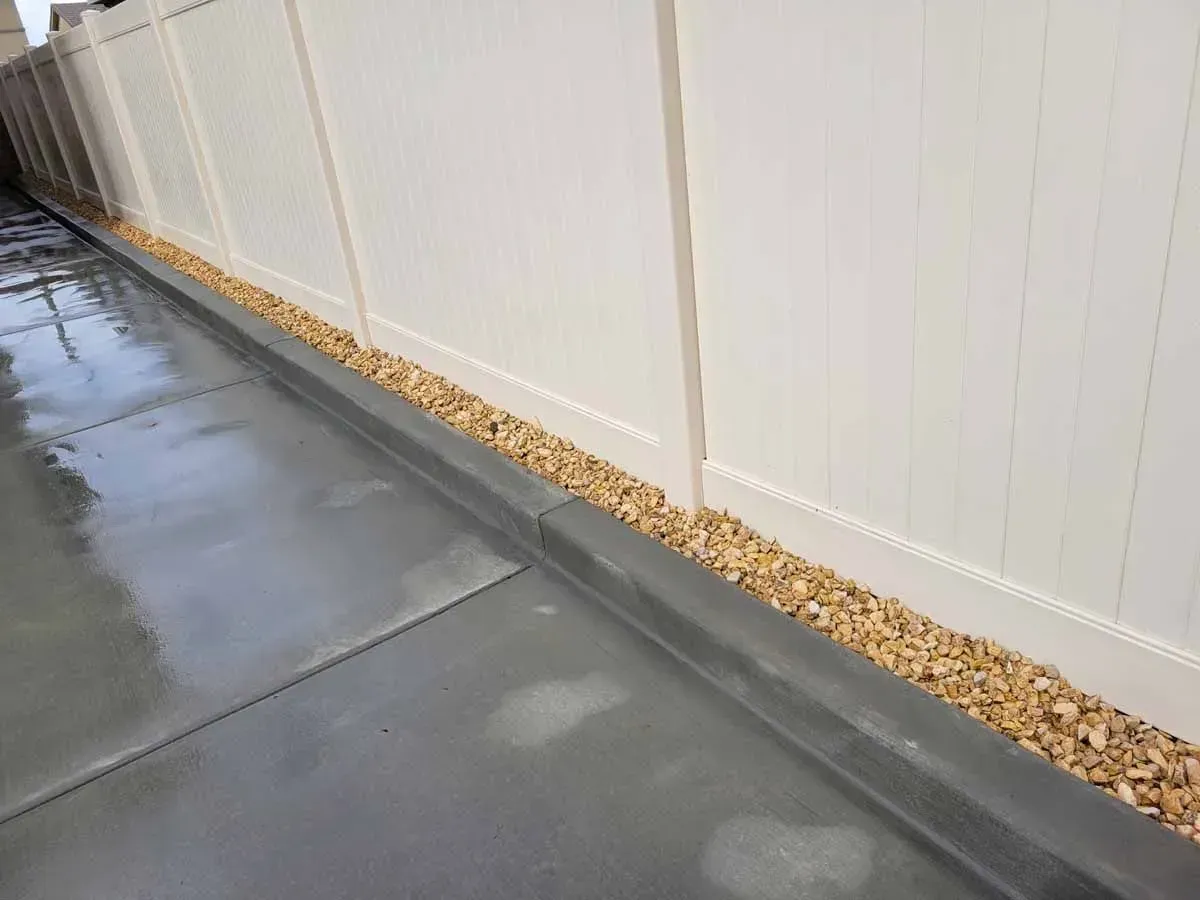 Gray concrete driveway with a white fence and tan gravel bed.