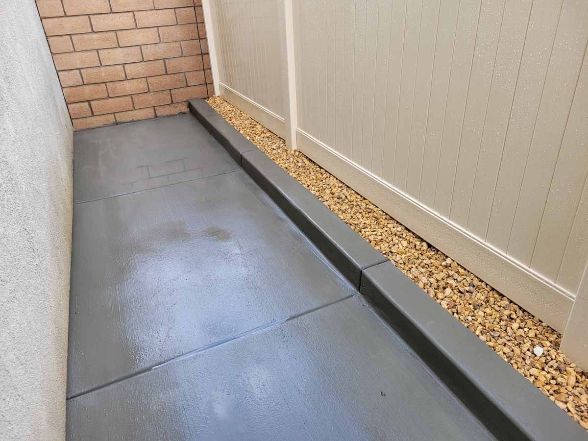 Narrow outdoor space with painted concrete, bordering gravel and a white fence.