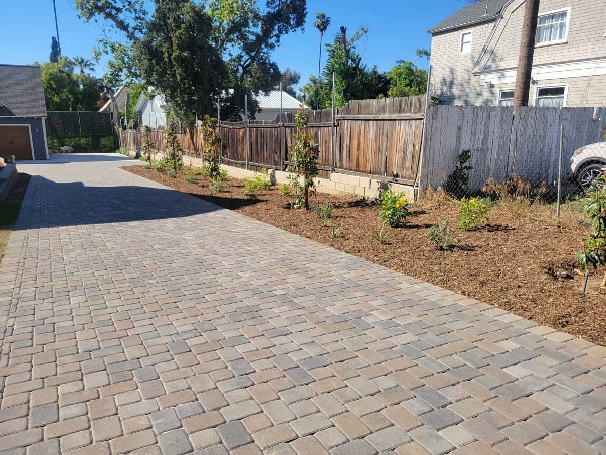 Brick driveway with a landscaped border next to a wooden fence and trees.