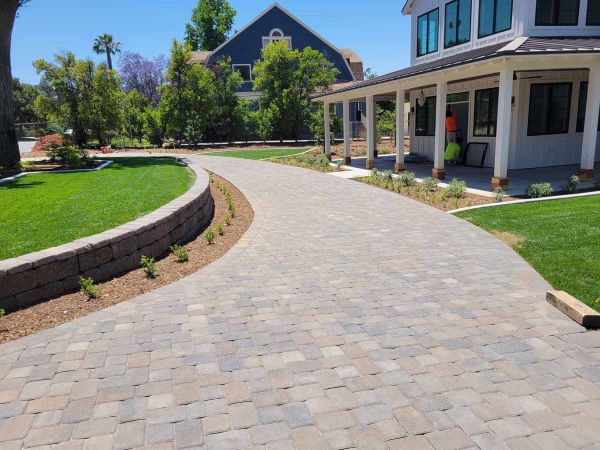 Paver driveway curves toward a house with a porch, bordered by grass and a low retaining wall.